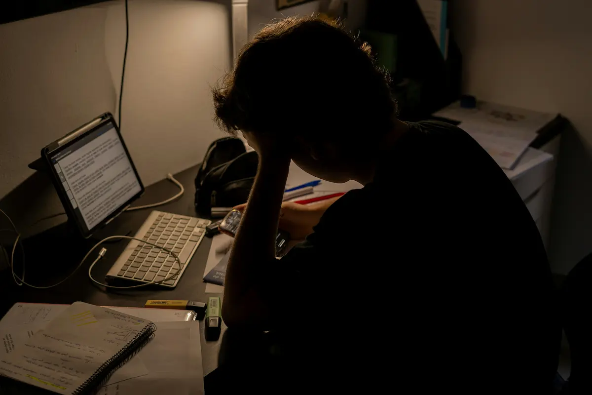 A person sitting at a cluttered desk in a dim room, head resting in their hand